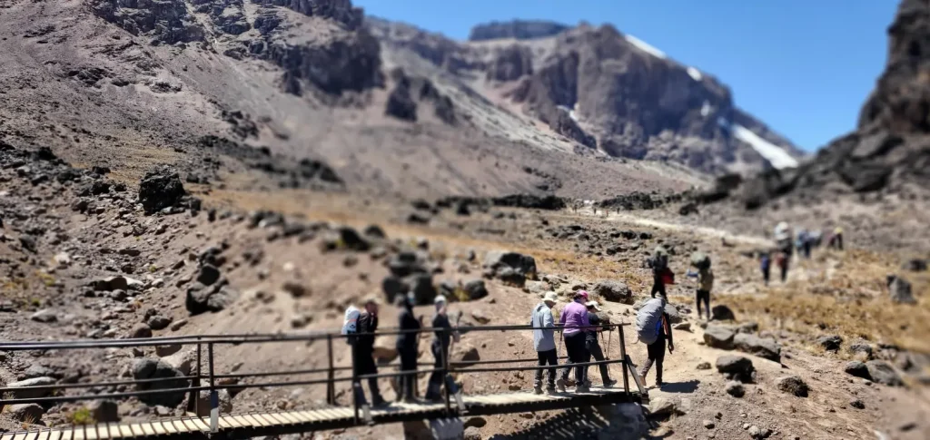 Group of trekkers crossing a wooden bridge on Mount Kilimanjaro during their ascent, surrounded by rocky terrain and clear blue skies.