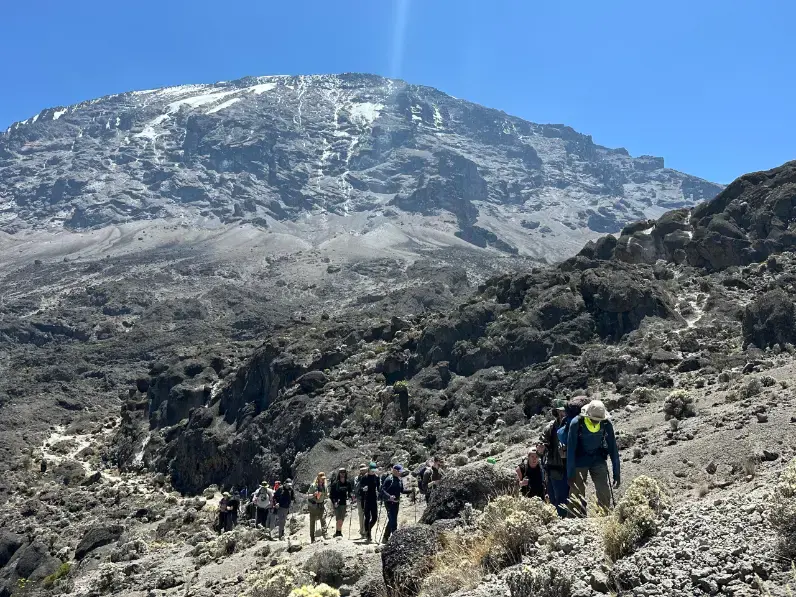 Trekkers hiking up the rocky slopes of Mount Kilimanjaro under a clear blue sky, with the snow-capped summit seen in the background, Africa’s tallest mountain at 5,895 meters.