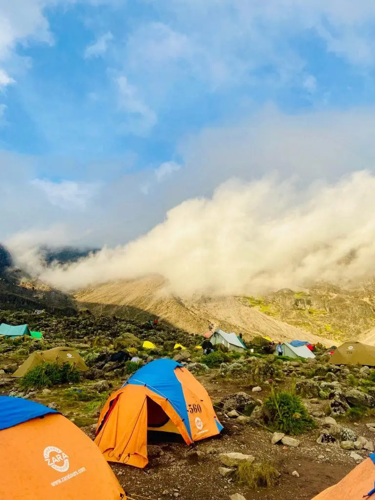 Colorful camping tents at a Kilimanjaro base camp surrounded by rocky terrain and mist-covered mountains under a bright blue sky.