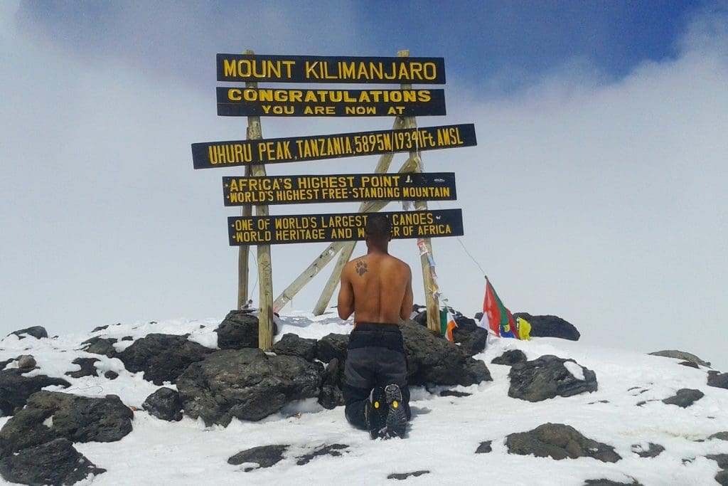 A climber kneels shirtless at the Uhuru Peak sign on Mount Kilimanjaro, surrounded by snow and congratulatory messages marking Africa’s highest point.