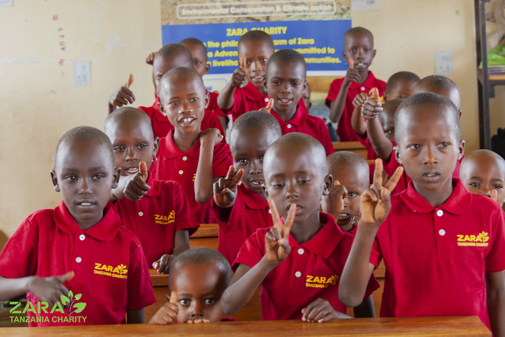 Young students in Zara Charity uniforms smiling and showing peace signs inside a classroom in Tanzania, part of Zara Tanzania Adventures’ education support initiative.
