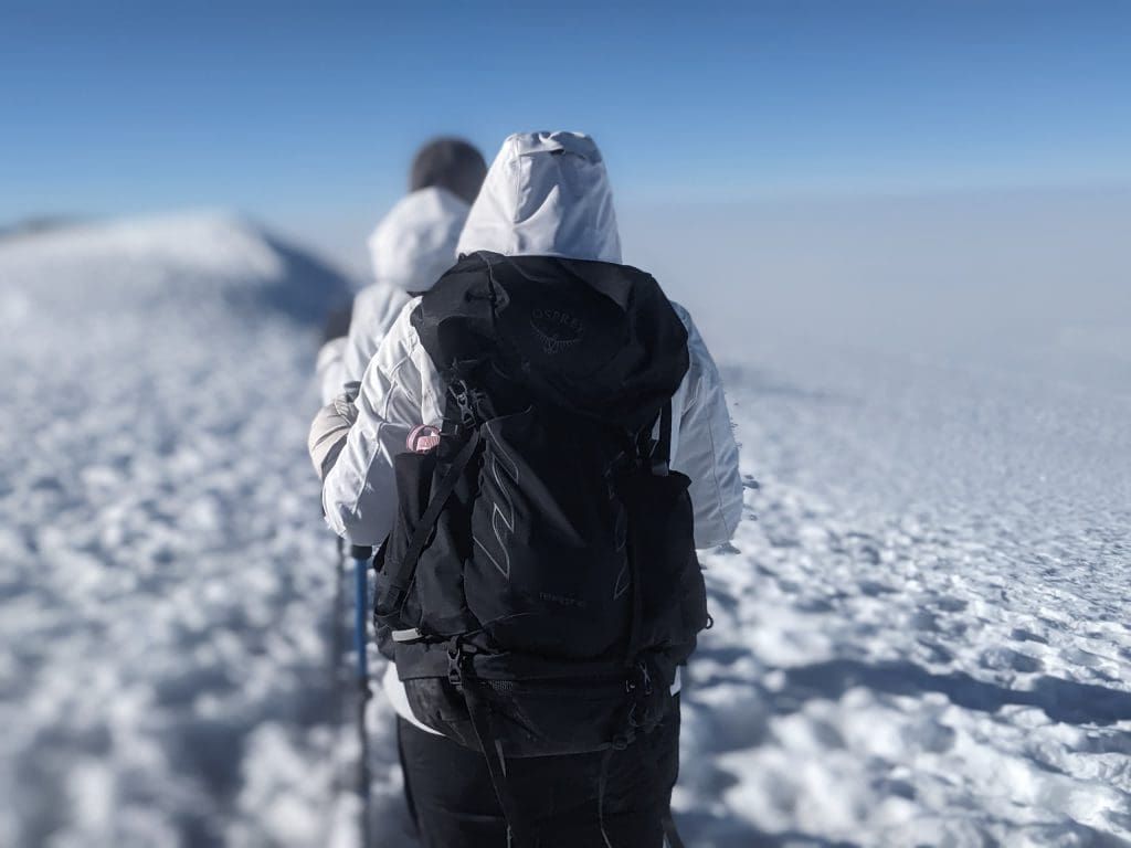 Climbers wearing winter gear and backpacks trek through the snowy summit of Mount Kilimanjaro under a clear blue sky.
