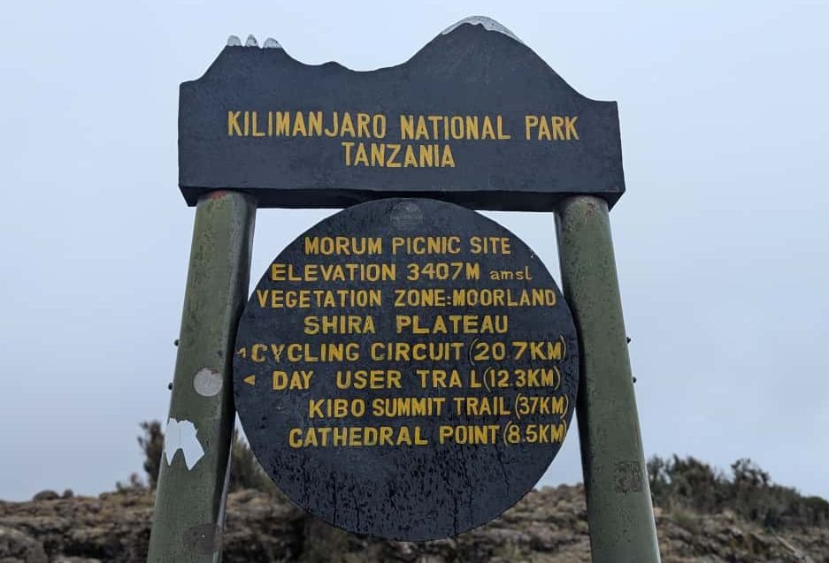 A signpost at morum picnic site in Kilimanjaro National Park showing the elevation of 3,407 meters and route directions to Kibo Summit Trail, Cathedral Point, and other trails.