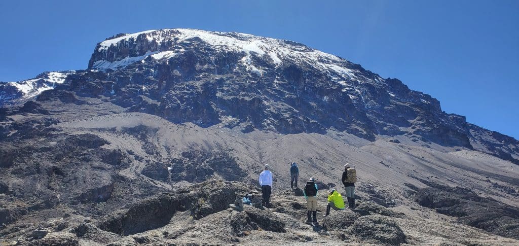 Climbers standing and resting on a rocky slope on Mount Kilimanjaro with the snow-capped summit rising above them under a clear blue sky.