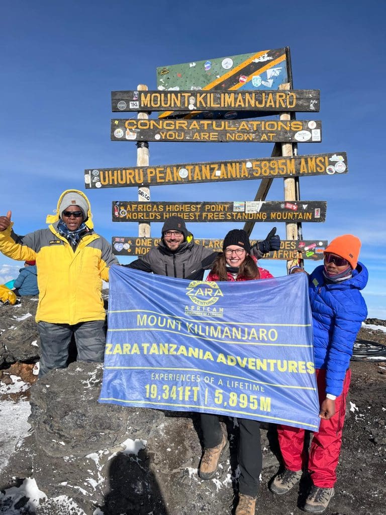 Climbers and guides celebrating at Uhuru Peak holding a Zara Tanzania Adventures banner in front of the Mount Kilimanjaro summit sign (5,895 m).