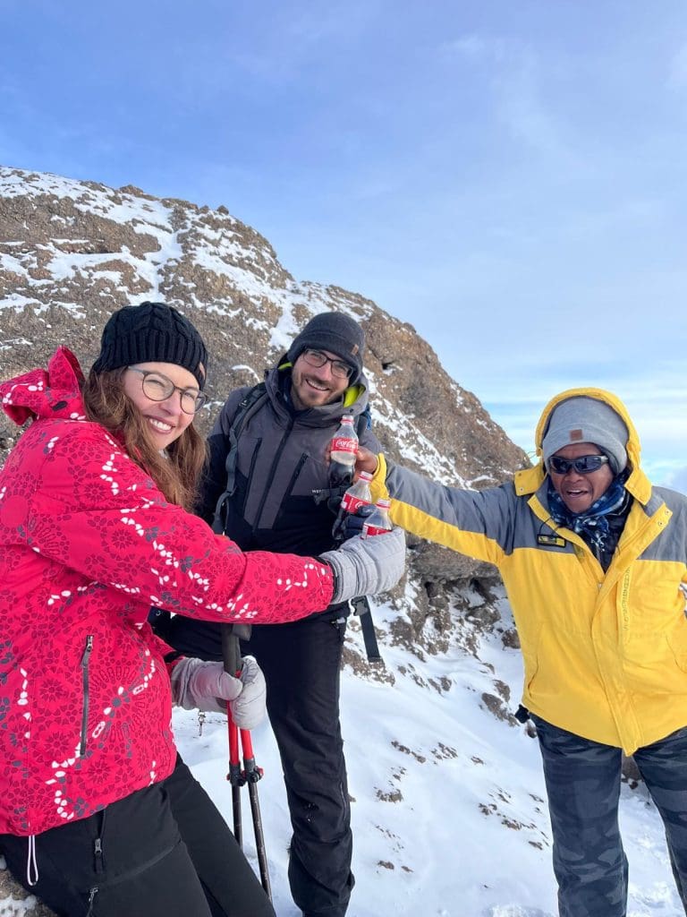 Climbers and their guide pausing on the snowy slopes of Mount Kilimanjaro, celebrating with drinks during the final ascent toward Uhuru Peak.